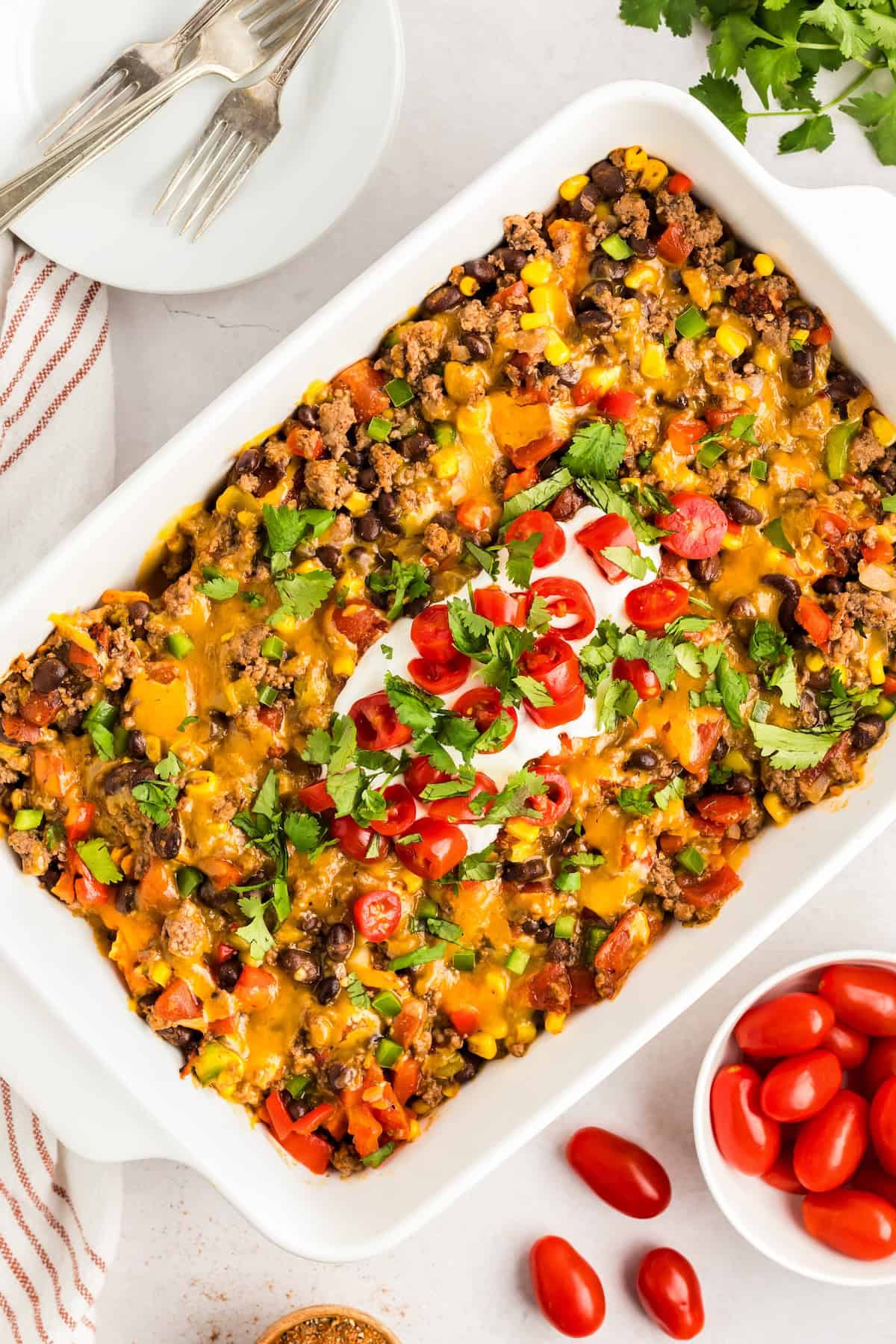 Crunchy, cheesy, beefy, and loaded with bold taco flavors, this Dorito Casserole recipe is quick to assemble and downright addictive! A baked taco casserole with Doritos in a white dish, topped with melted cheese, chopped tomatoes, cilantro, and a dollop of sour cream.
