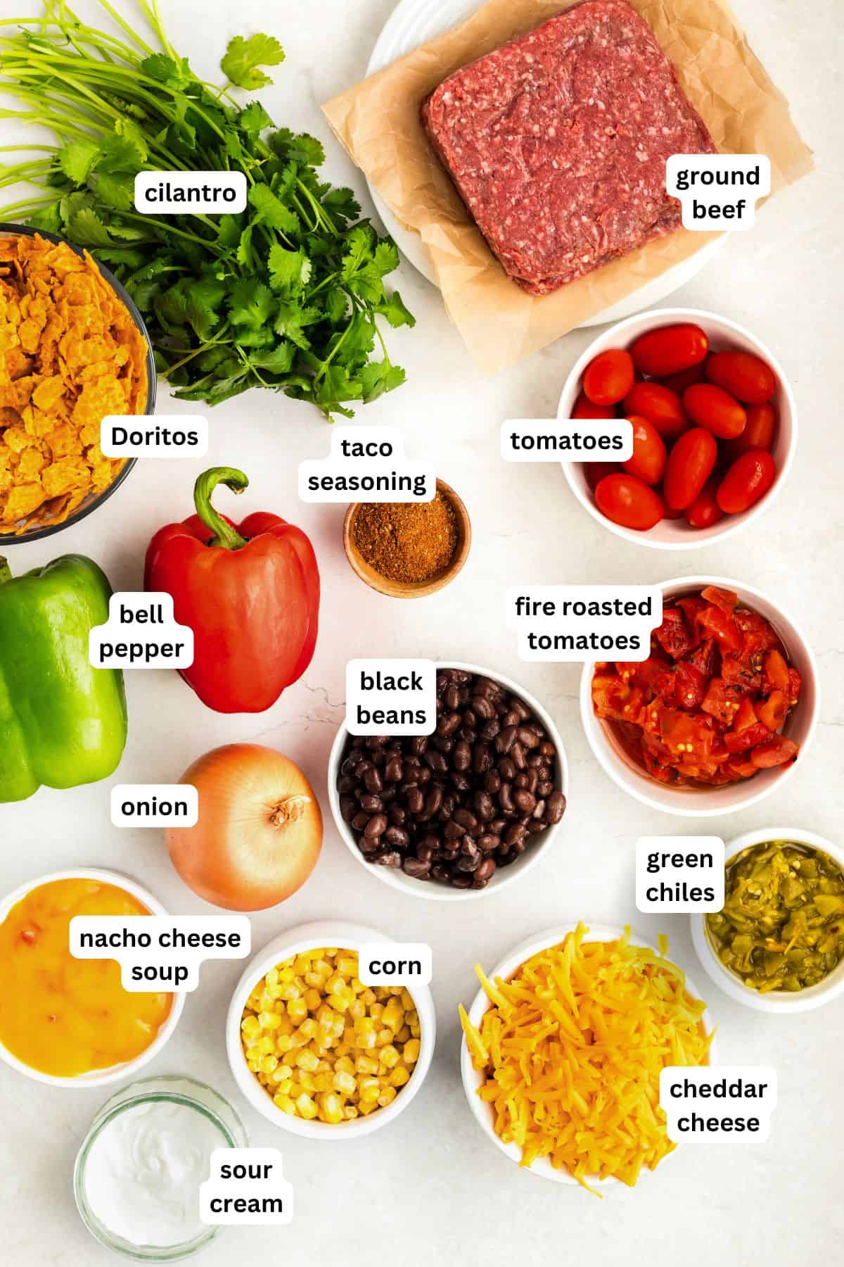 Crunchy, cheesy, beefy, and loaded with bold taco flavors, this Dorito Casserole recipe is quick to assemble and downright addictive! Ingredients for a Dorito casserole recipe arranged on a white surface: ground beef, cilantro, Doritos, taco seasoning, tomatoes, bell peppers, onion, fire roasted tomatoes, black beans, green chiles, nacho cheese soup, corn, cheddar cheese, and sour cream.