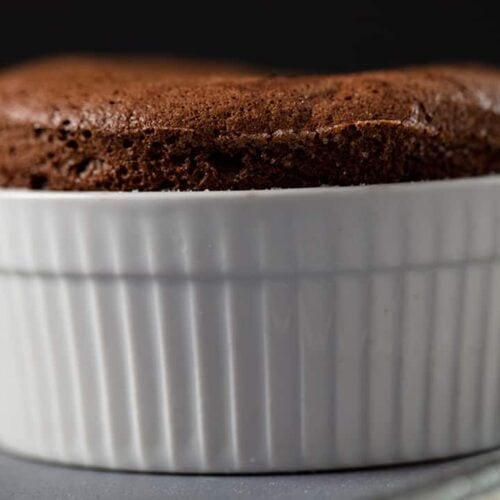A chocolate soufflé rises above the rim of a white, ribbed ramekin, sitting on a gray surface with a spoon nearby against a dark background.