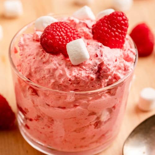 A glass cup filled with a fluffy pink dessert, topped with raspberries and mini marshmallows, sits on a wooden surface with a spoon and scattered marshmallows and raspberries nearby.