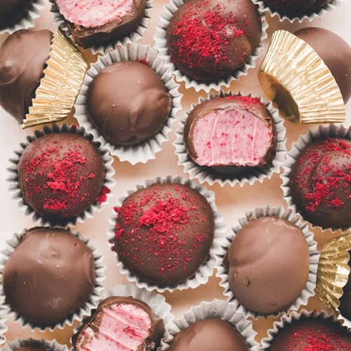 Chocolate raspberry truffles arranged on a tray, some with red raspberry crumbs on top, some with bites taken out to reveal a pink raspberry filling inside, and a few placed in gold foil wrappers.