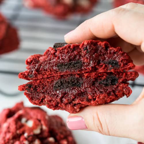 A hand with pink nail polish holds a red velvet cookie broken in half, revealing a dark chocolate or cookie filling inside. More cookies and a cooling rack are visible in the background.