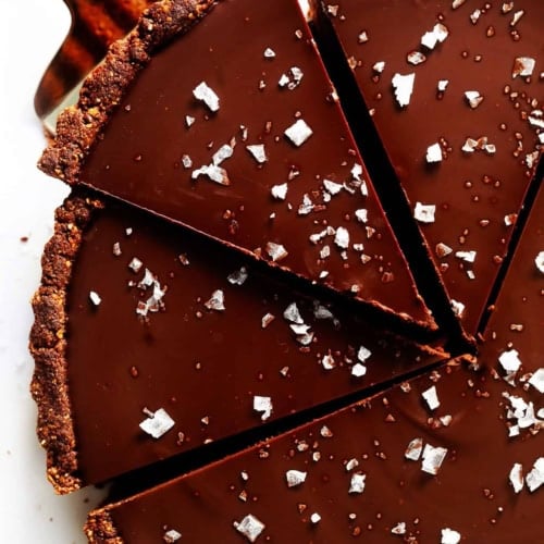 A hand uses a pie server to lift a slice from a rich chocolate tart topped with flaky sea salt; the tart is cut into five large slices on a white background.