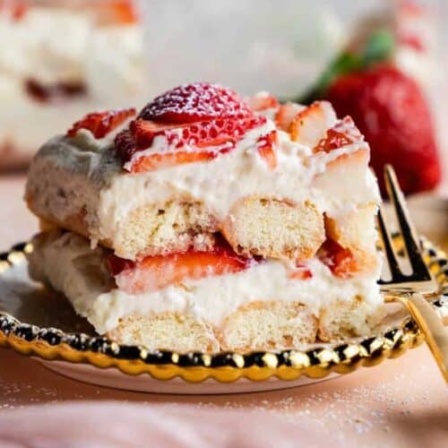 A close-up of a slice of strawberry tiramisu on a decorative plate with a fork, showing layers of ladyfingers, creamy filling, and fresh strawberries. Whole strawberries and ladyfingers are in the background.
