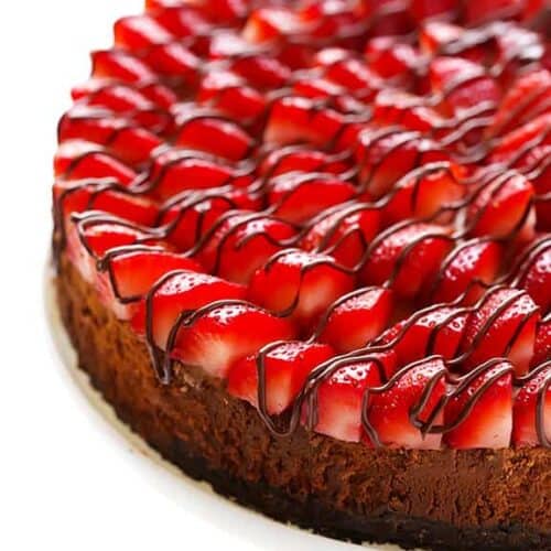 A cheesecake topped with glossy, sliced strawberries arranged in layers and drizzled with chocolate, displayed on a white cake stand against a white background.