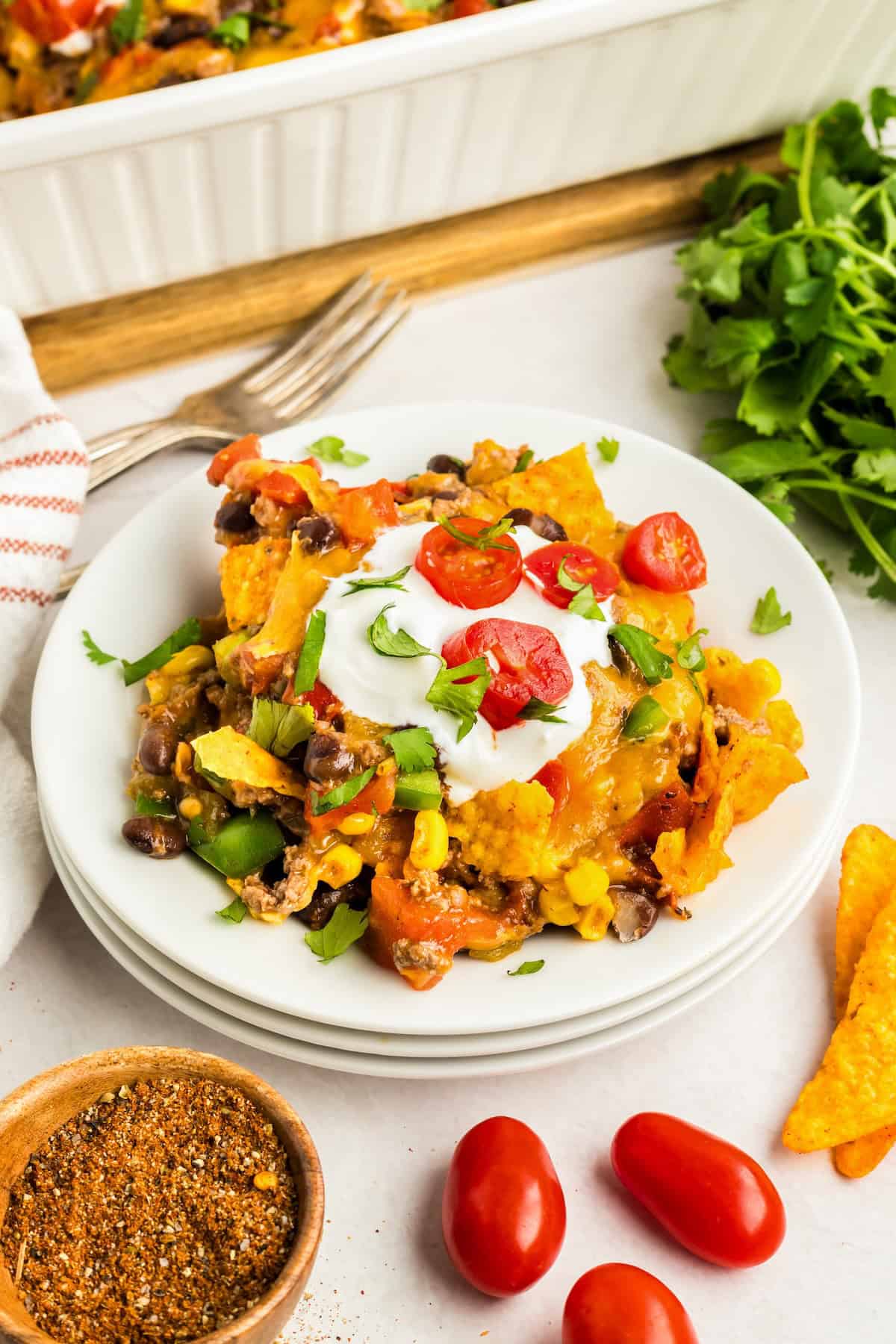 Crunchy, cheesy, beefy, and loaded with bold taco flavors, this Dorito Casserole recipe is quick to assemble and downright addictive! A serving of taco Dorito casserole topped with sour cream, chopped cilantro, and sliced cherry tomatoes is on a white plate. Ingredients like tortilla chips, seasoning, and cherry tomatoes are nearby.