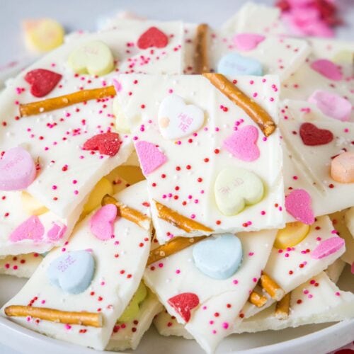 A plate of white chocolate bark topped with colorful heart-shaped candies, pretzel sticks, and red sprinkles, arranged in overlapping pieces.