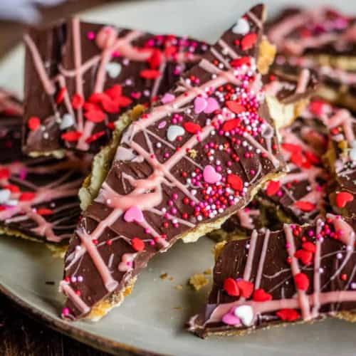 A plate of Valentine’s Day Saltine Cracker Toffee (Cracker Candy) topped with pink and red heart-shaped sprinkles and drizzled with pink and white icing, arranged on a white plate.