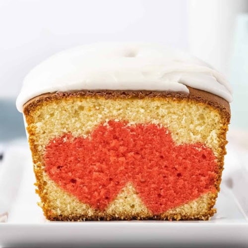 A slice of vanilla heart surprise cake with white icing, featuring a red heart shape baked into the center, displayed on a white plate.