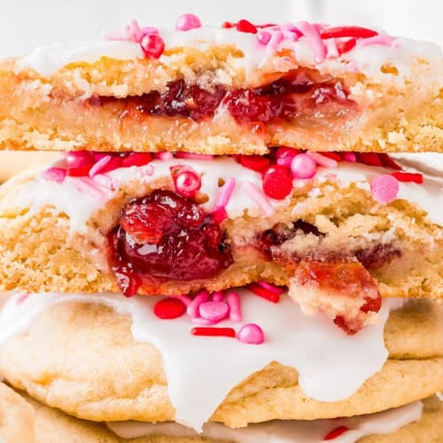 A close-up of thick, frosted sugar cookies filled with cherry jam. The cookies are topped with white icing and pink and red sprinkles, with one cookie cut in half to show the gooey cherry filling inside.