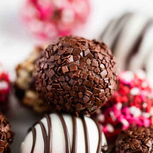 A close-up of assorted chocolate truffles, including one with chocolate sprinkles, one with white chocolate and dark chocolate drizzle, and others decorated with pink, red, and white sprinkles in the background.
