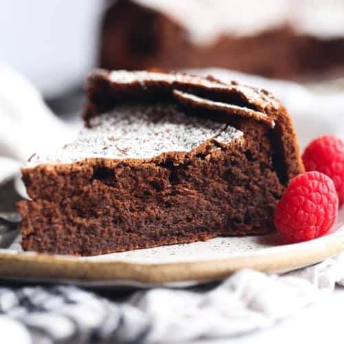 A slice of rich chocolate cake topped with powdered sugar sits on a plate, with two fresh raspberries beside it. The background is softly blurred, highlighting the dessert.