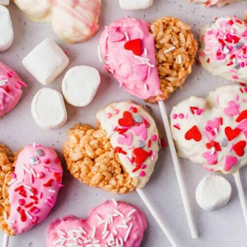 Heart-shaped Rice Krispie treats on sticks, decorated with pink and white chocolate and festive sprinkles, surrounded by marshmallows on a light background.