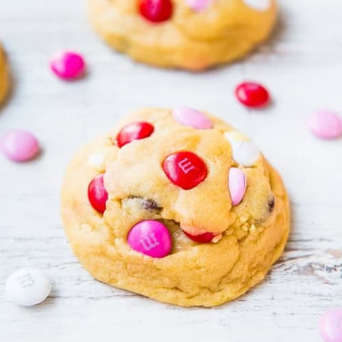 A soft, thick cookie topped with red, white, and pink candy-coated chocolates sits on a light wooden surface, surrounded by a few scattered candies.