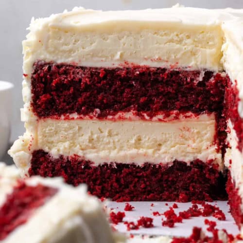 A close-up of a sliced red velvet cake cheesecake with two layers of cheesecake and creamy white frosting, showing the rich red and creamy white layers inside.