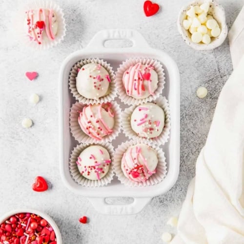 A white dish holds six pink and white cake balls with festive sprinkles. Around it are bowls of white chocolate chips and red sprinkles, a bottle of milk, and red heart decorations on a light surface.