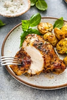 A plate with a juicy, seasoned baked chicken breast, sliced with a fork, served alongside roasted baby potatoes and garnished with fresh basil leaves.