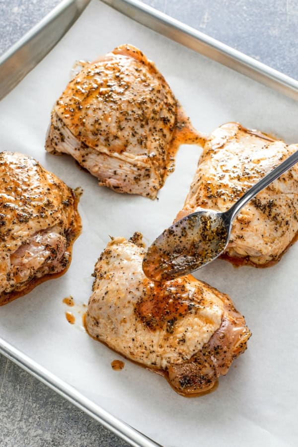Oh-so-juicy with perfectly crisp skin, this Oven Baked Chicken recipe marinates chicken breast or thighs with a garlicky, citrus glaze. Four seasoned raw chicken thighs on a parchment-lined baking sheet, with a spoon drizzling marinade over one of the thighs.