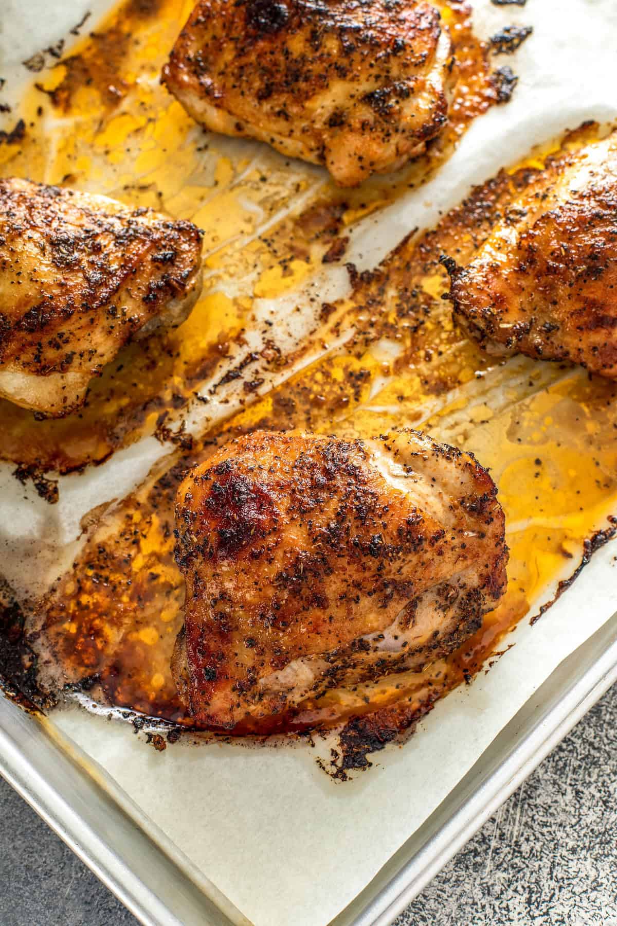 Oh-so-juicy with perfectly crisp skin, this Oven Baked Chicken recipe marinates chicken breast or thighs with a garlicky, citrus glaze. Four oven baked chicken thighs resting on a parchment-lined baking sheet, with golden-brown skin and juices pooling around each piece.