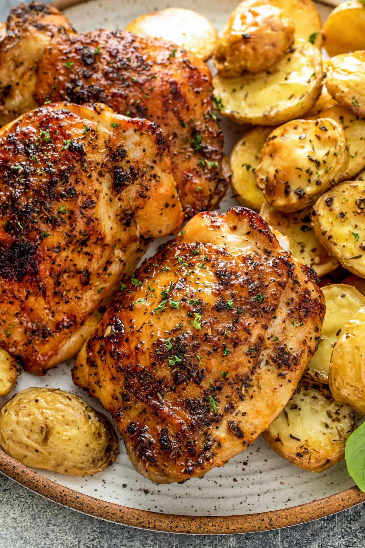 Oh-so-juicy with perfectly crisp skin, this Oven Baked Chicken recipe marinates chicken breast or thighs with a garlicky, citrus glaze. A plate with three pieces of roasted, seasoned chicken and slices of golden, herb-roasted potatoes, garnished with fresh herbs.