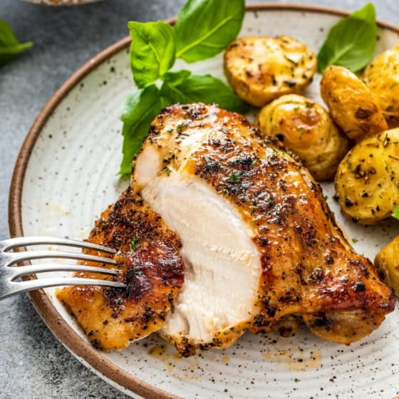 A plate with a juicy, seasoned baked chicken breast, sliced with a fork, served alongside roasted baby potatoes and garnished with fresh basil leaves.