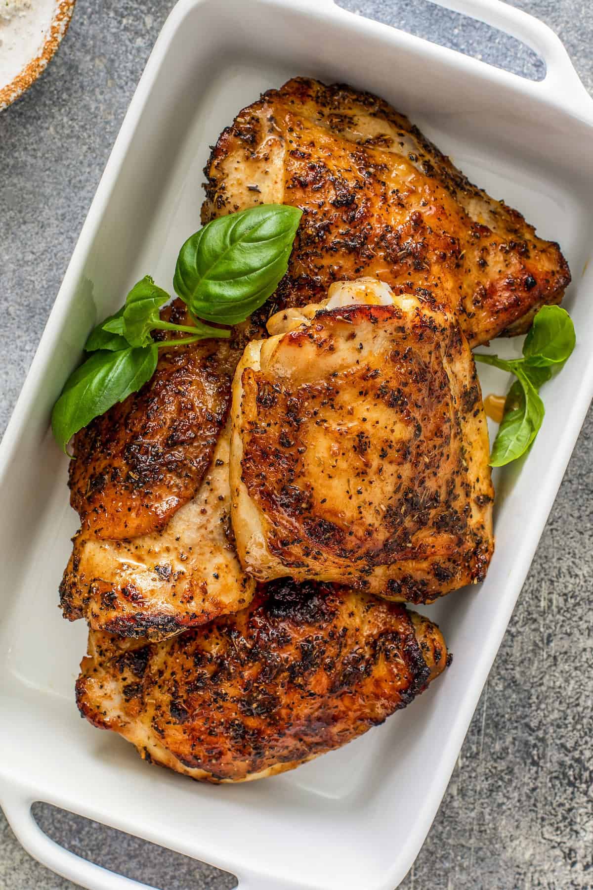 Oh-so-juicy with perfectly crisp skin, this Oven Baked Chicken recipe marinates chicken breast or thighs with a garlicky, citrus glaze. Three pieces of seasoned and baked chicken breasts and thighs in a white rectangular dish, garnished with fresh basil leaves, on a gray surface.