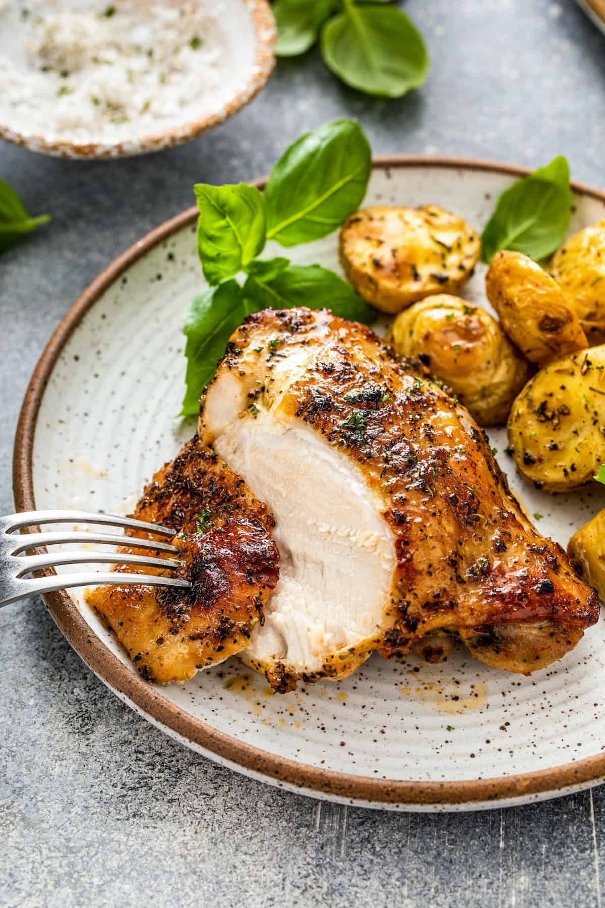 Oh-so-juicy with perfectly crisp skin, this Oven Baked Chicken recipe marinates chicken breast or thighs with a garlicky, citrus glaze. A plate with a juicy, seasoned baked chicken breast, sliced with a fork, served alongside roasted baby potatoes and garnished with fresh basil leaves.
