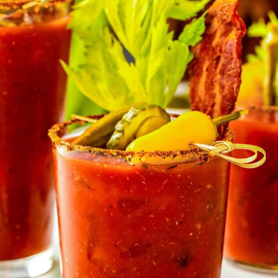 The best Bloody Mary drink served in a glass garnished with celery, crispy bacon, pickles, olives, and a yellow tomato, with a seasoned rim. Two more similar drinks are visible in the background.