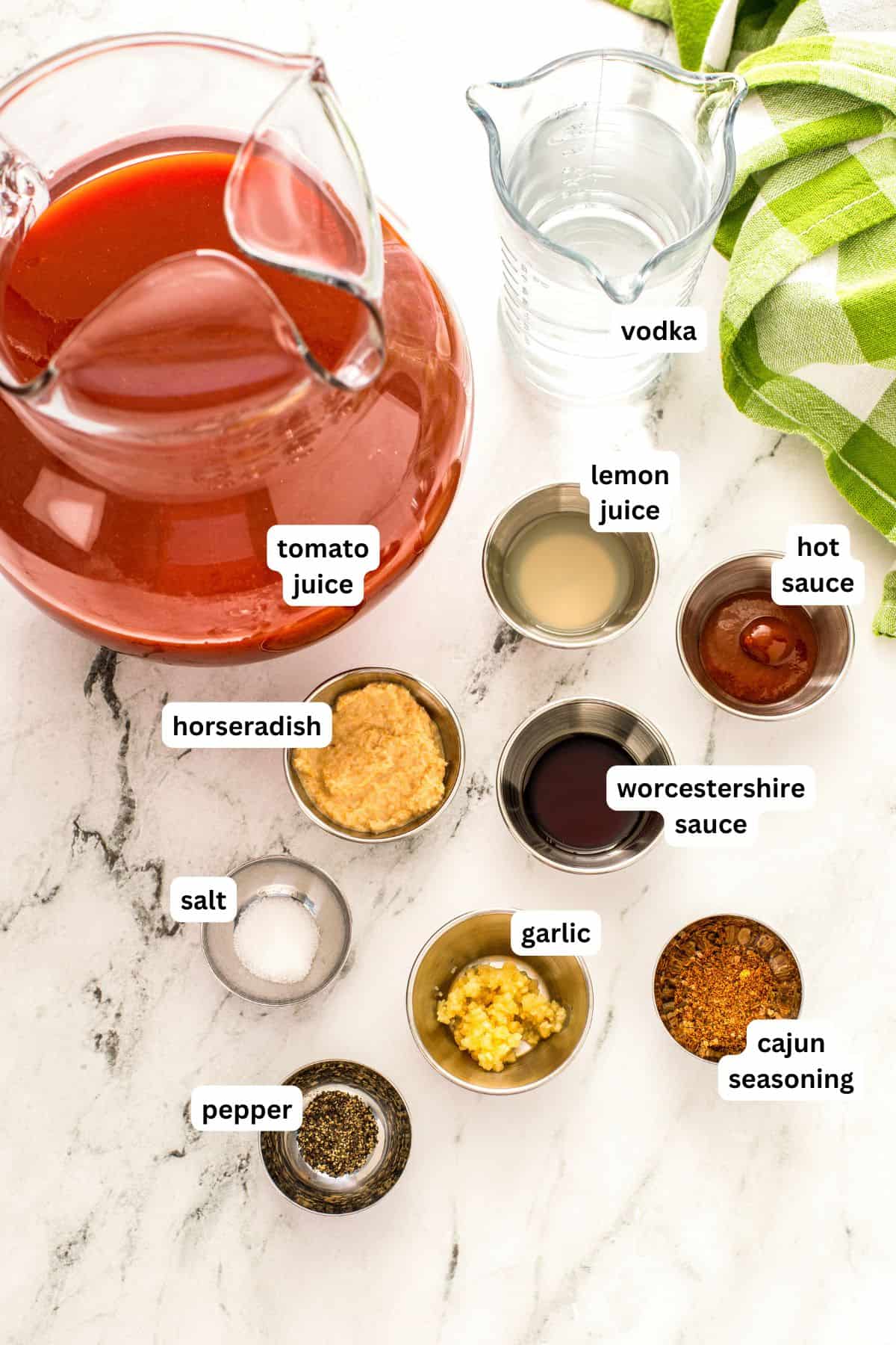 Packed with bold Cajun seasoning, zippy horseradish, and fresh garlic for just the right amount of heat, this is the best Bloody Mary recipe! Overhead view of labeled Bloody Mary ingredients on a marble counter: pitcher of tomato juice, jar of vodka, bowls of lemon juice, hot sauce, Worcestershire sauce, horseradish, salt, pepper, garlic, and Cajun seasoning.