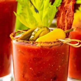 A glass of Bloody Mary vodka cocktail garnished with celery, bacon, pickles, and a yellow pepper, with a spiced rim.