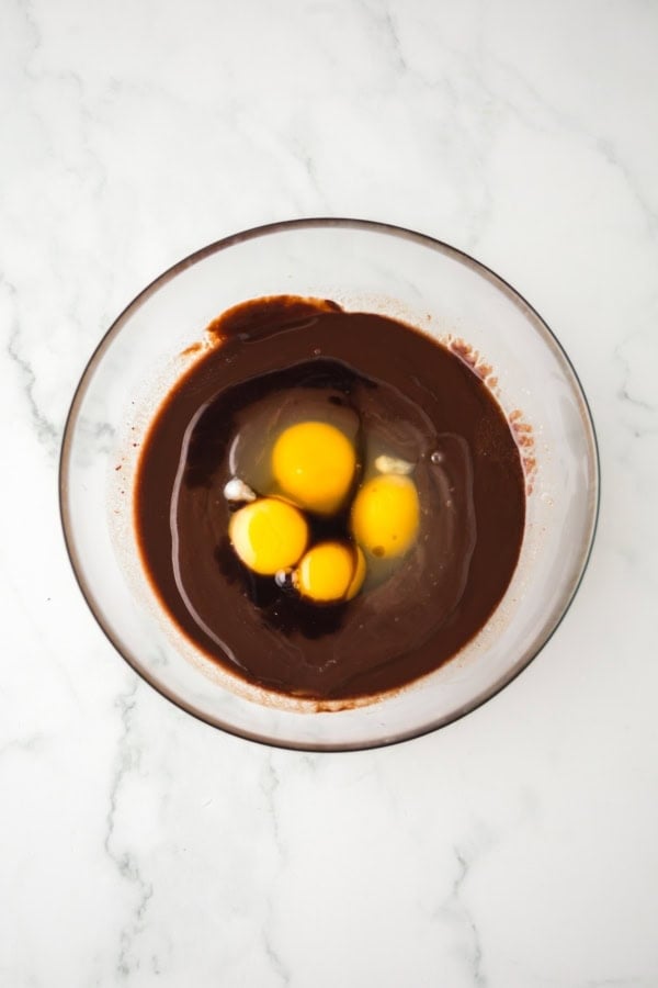 A glass bowl on a marble surface containing melted chocolate and butter mixture with raw eggs on top, not yet mixed together.