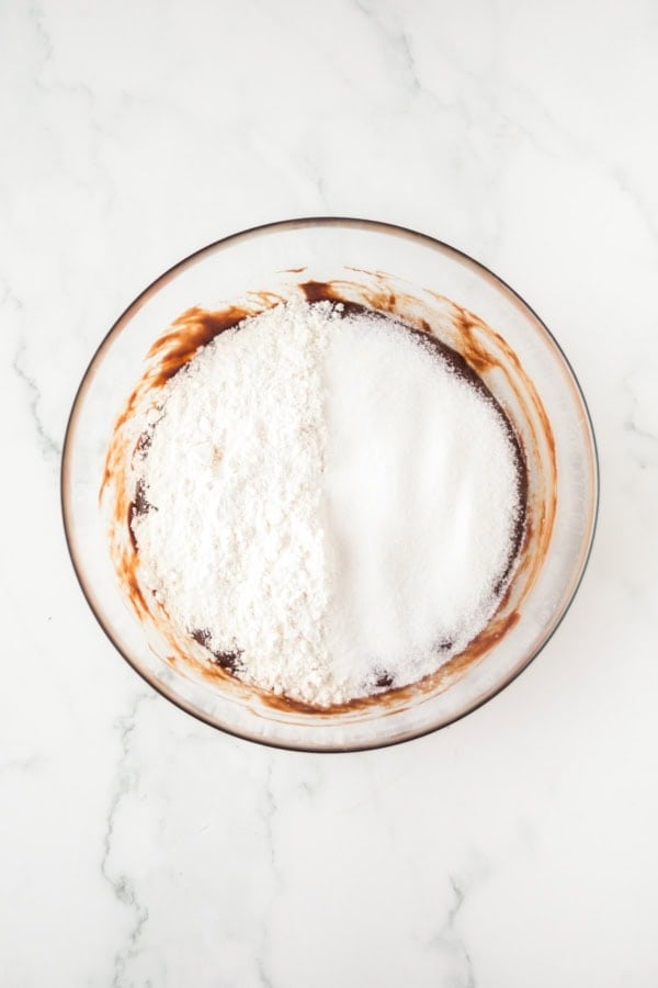 A glass bowl containing chocolate mixture, half covered with a heap of white flour and sugar, sits on a marble countertop.