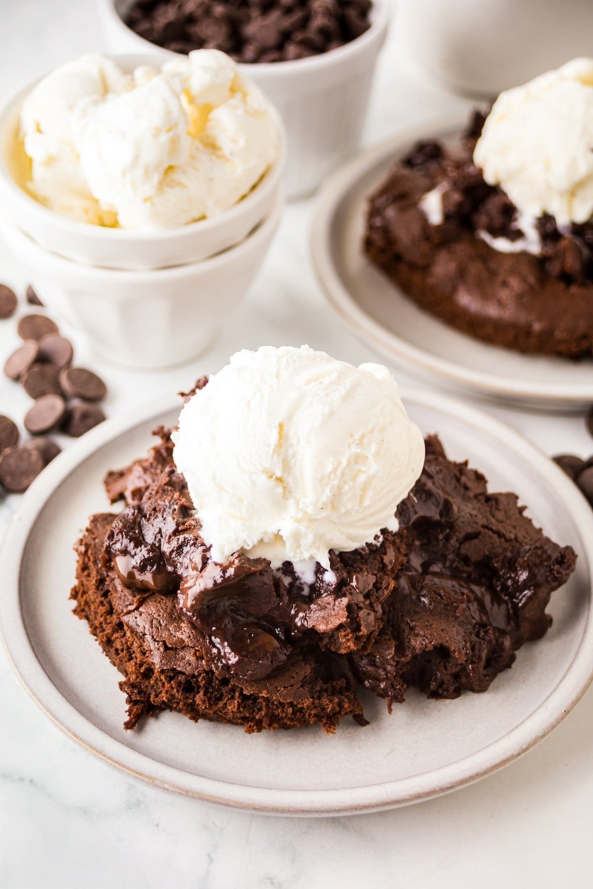 A close-up of a chocolate pudding cake topped with a scoop of vanilla ice cream on a white plate, with chocolate chips scattered nearby and a bowl of ice cream in the background.