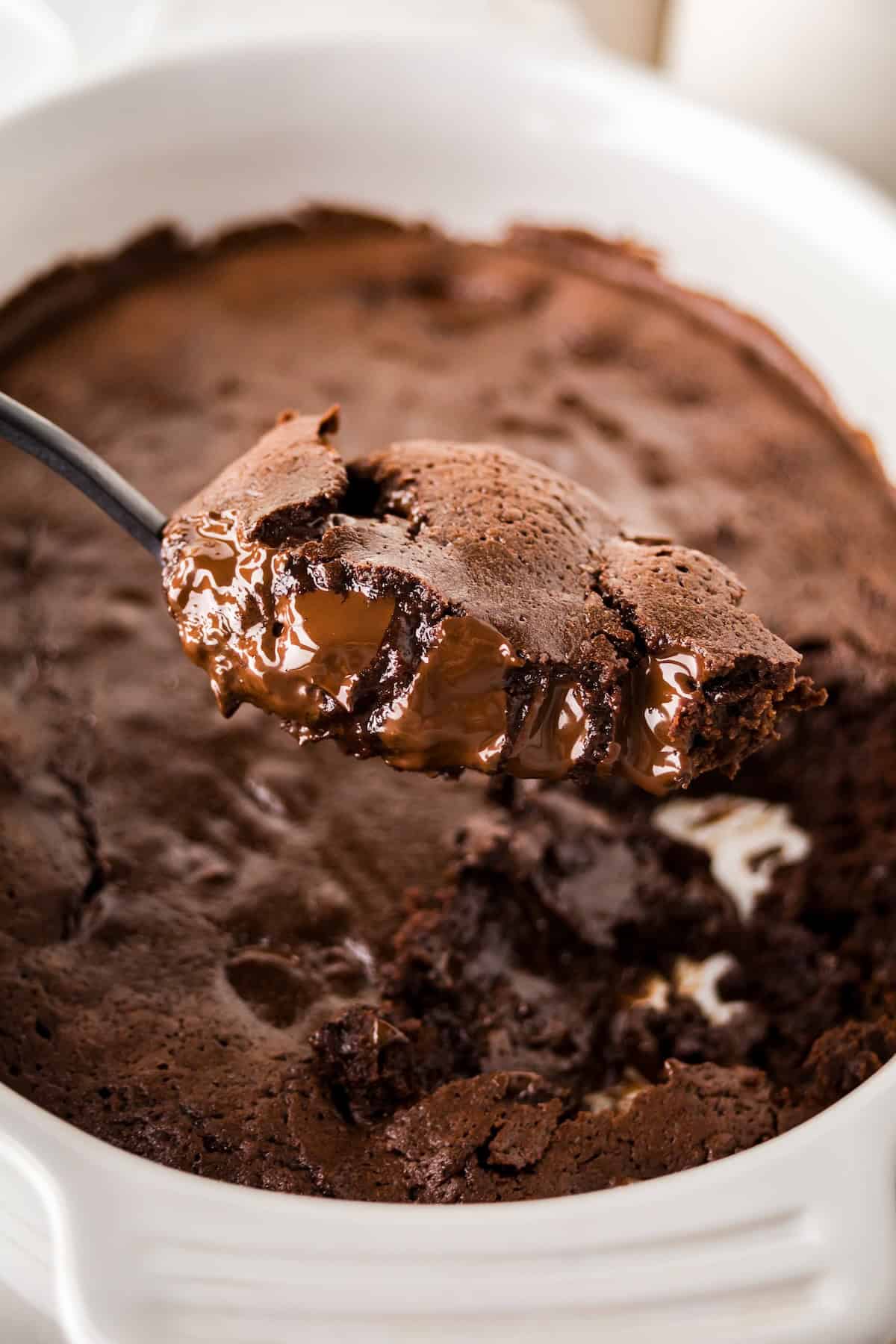 A close-up of a spoon lifting a serving of gooey chocolate pudding cake from a white dish, showing the rich, fudgy interior and crispy edges.