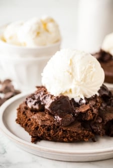 A decadent chocolate pudding cake topped with a scoop of vanilla ice cream sits on a plate. The cake appears gooey and rich, with a melted chocolate pudding filling visible.