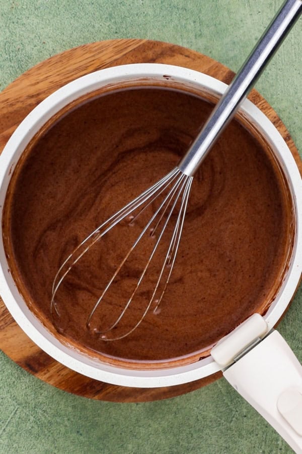 Rich, dark, and unbelievably moist, this Guinness Chocolate Cake is frosted with a sweet Bailey's buttercream. Perfect for St. Patrick's Day! A metal whisk rests in a saucepan filled with a smooth melted chocolate mixture, placed on a round wooden trivet.