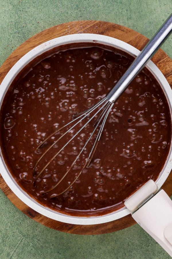 Rich, dark, and unbelievably moist, this Guinness Chocolate Cake is frosted with a sweet Bailey's buttercream. Perfect for St. Patrick's Day! A bowl filled with chocolate cake batter sits on a wooden board. A metal whisk rests in the batter, which has small bubbles on its surface.