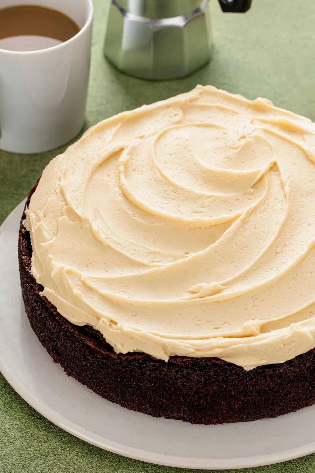Rich, dark, and unbelievably moist, this Guinness Chocolate Cake is frosted with a sweet Bailey's buttercream. Perfect for St. Patrick's Day! A Guinness chocolate cake topped with swirls of Baileys buttercream frosting sits on a white plate next to a cup of coffee and a metal coffee pot.