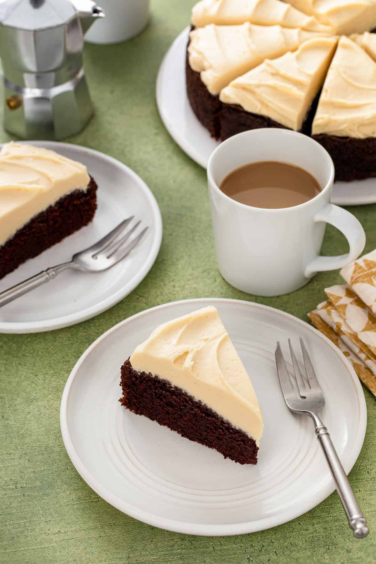 Rich, dark, and unbelievably moist, this Guinness Chocolate Cake is frosted with a sweet Bailey's buttercream. Perfect for St. Patrick's Day! A slice of rich and moist Guinness chocolate cake topped with Baileys buttercream frosting on a white plate with a fork, next to a mug of coffee. The rest of the cake and another slice are in the background.