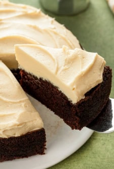 A slice of rich Guinness chocolate cake with creamy Baileys Irish Cream frosting is being lifted with a cake server off a white cake plate.