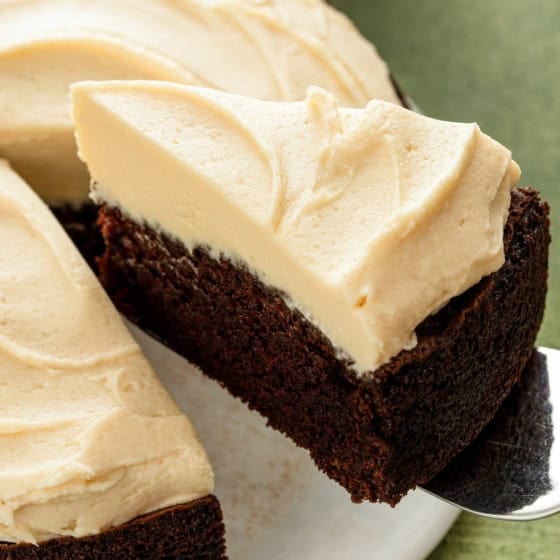 A slice of rich Guinness chocolate cake with creamy Baileys Irish Cream frosting is being lifted with a cake server off a white cake plate.