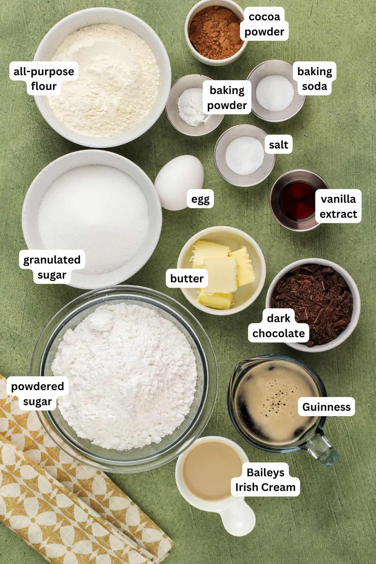 Rich, dark, and unbelievably moist, this Guinness Chocolate Cake is frosted with a sweet Bailey's buttercream. Perfect for St. Patrick's Day! Overhead view of labeled ingredients for Guinness chocolate cake recipe, including flour, cocoa powder, baking powder, baking soda, salt, vanilla, egg, sugar, butter, dark chocolate, Guinness, Baileys Irish Cream, and powdered sugar.