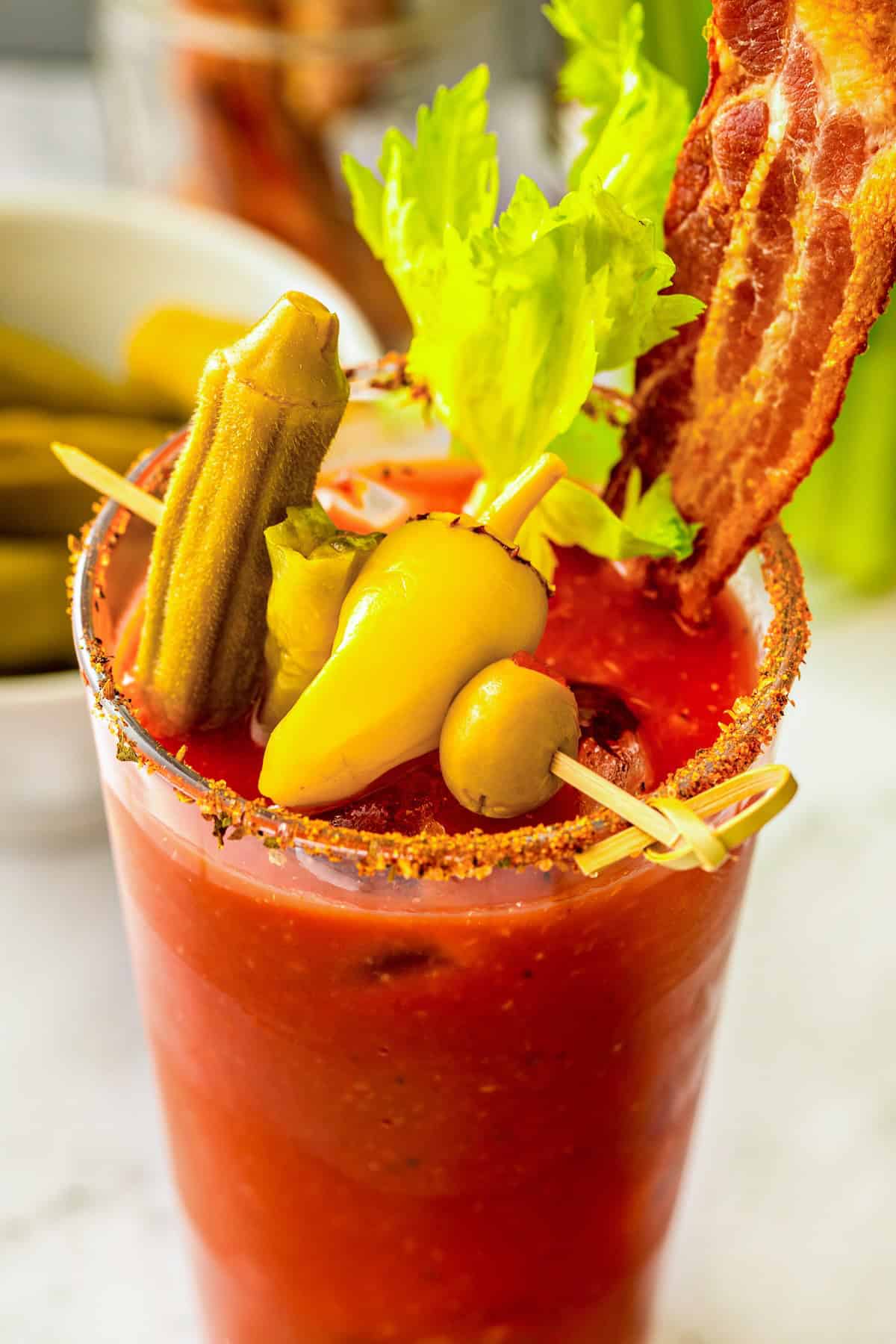 Packed with bold Cajun seasoning, zippy horseradish, and fresh garlic for just the right amount of heat, this is the best Bloody Mary recipe! A loaded Bloody Mary cocktail garnished with a celery stalk, pickled okra, a yellow pepper, a green olive, and a crispy bacon strip, with a seasoned salt rim on the glass.