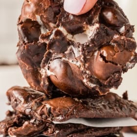 A hand holds a gooey, chocolate fudge cookie with melted marshmallows over a stack of similar chocolate cookies.