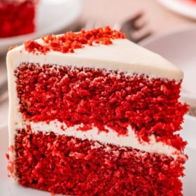 A slice of the best red velvet cake with cream cheese frosting on a plate, showing two layers of red cake with creamy filling, with more slices in the background.