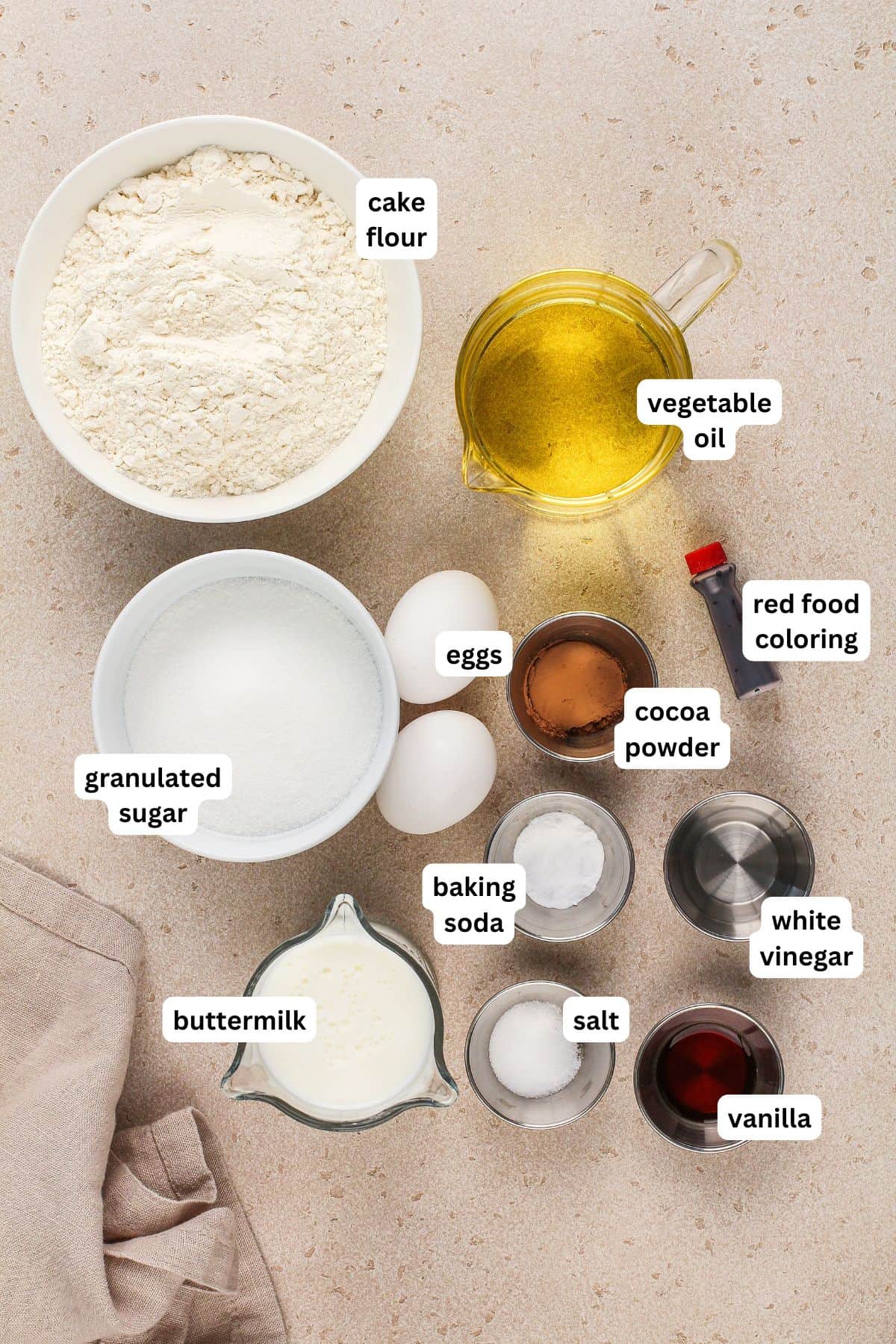 This is the best Red Velvet Cake recipe with a moist, soft crumb and a classic tangy flavor from buttermilk and sweet cream cheese frosting! A top-down view of labeled baking ingredients for a homemade red velvet cake recipe on a counter, including cake flour, vegetable oil, red food coloring, eggs, granulated sugar, cocoa powder, baking soda, white vinegar, salt, buttermilk, and vanilla.