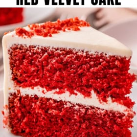 A close-up of a slice of homemade red velvet cake with two layers, creamy white frosting, and red cake crumbs on top, on a white plate.