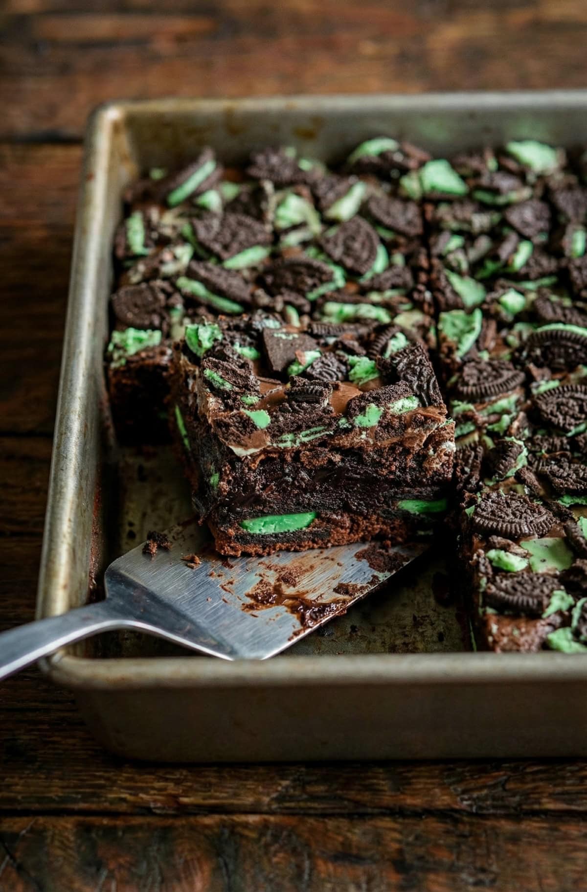 Fudgy chocolate brownies are filled with layers of Mint Oreos and Andes Mints to create these epic Andes Mint Brownies. A tray of mint chocolate brownies topped with crushed mint Oreo cookies and andes mints, with one brownie being lifted out by a metal spatula on a wooden surface.