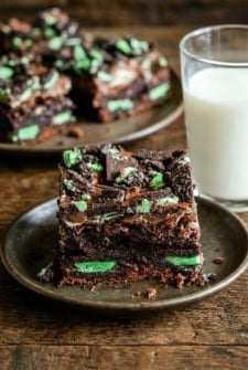 Chocolate mint brownies with Andes mints and mint Oreos on a plate, next to a glass of milk. More brownies are visible on a plate in the background.