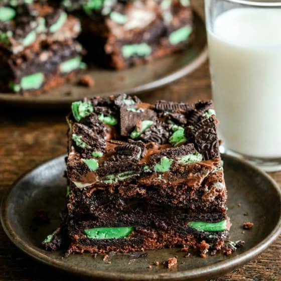 Chocolate mint brownies with Andes mints and mint Oreos on a plate, next to a glass of milk. More brownies are visible on a plate in the background.